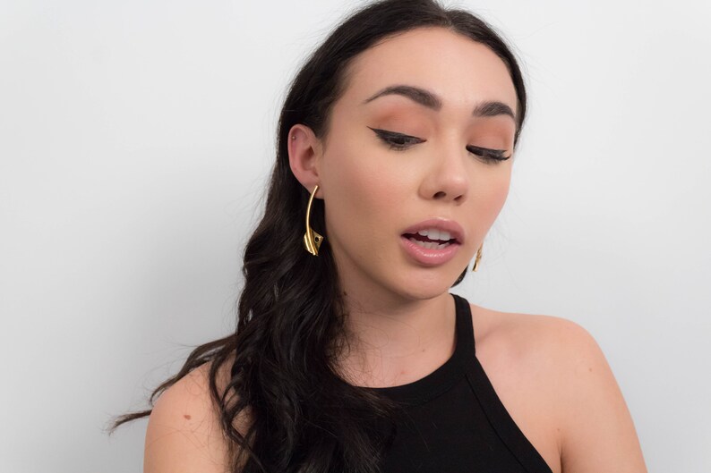 May include: A woman with long dark hair wearing gold hoop earrings. She is wearing a black tank top and has a neutral makeup look with winged eyeliner.