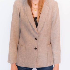 Puede incluir: Una chaqueta de traje de cuadros marrones y blancos con cuello de muesca y tres botones negros. La chaqueta tiene dos bolsillos de solapa y mangas largas.