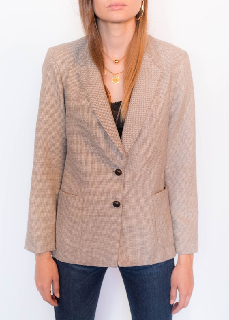 Puede incluir: Una chaqueta de traje de cuadros marrones y blancos con cuello de muesca y tres botones negros. La chaqueta tiene dos bolsillos de solapa y mangas largas.