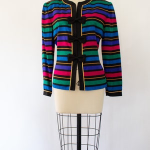 May include: A black and multi-colored striped blazer with gold trim and bow accents. The blazer has a long sleeve and a v-neck.