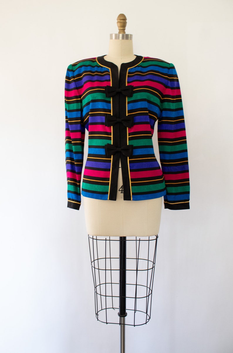 May include: A black and multi-colored striped blazer with gold trim and bow accents. The blazer has a long sleeve and a v-neck.