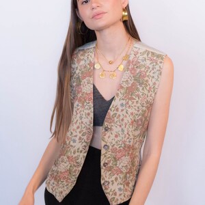 May include: A floral patterned vest with a button closure. The vest is beige with pink, green, and blue flowers. The model is wearing the vest over a black top and black pants.