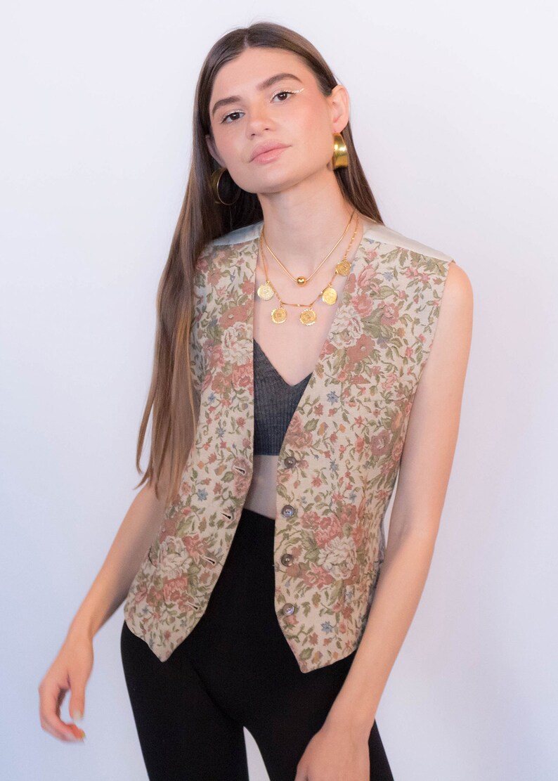 May include: A floral patterned vest with a button closure. The vest is beige with pink, green, and blue flowers. The model is wearing the vest over a black top and black pants.