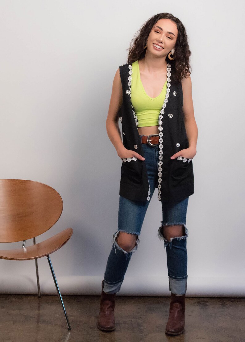 May include: A woman wearing a black vest with white floral trim, a green tank top, blue jeans with rips, and brown boots. She is smiling and has her hands in her pockets.