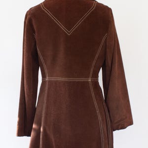 May include: A brown suede jacket with long sleeves and a tailored fit. The jacket features white stitching along the seams, collar, and a decorative V-shape on the back. The jacket is displayed on a mannequin.