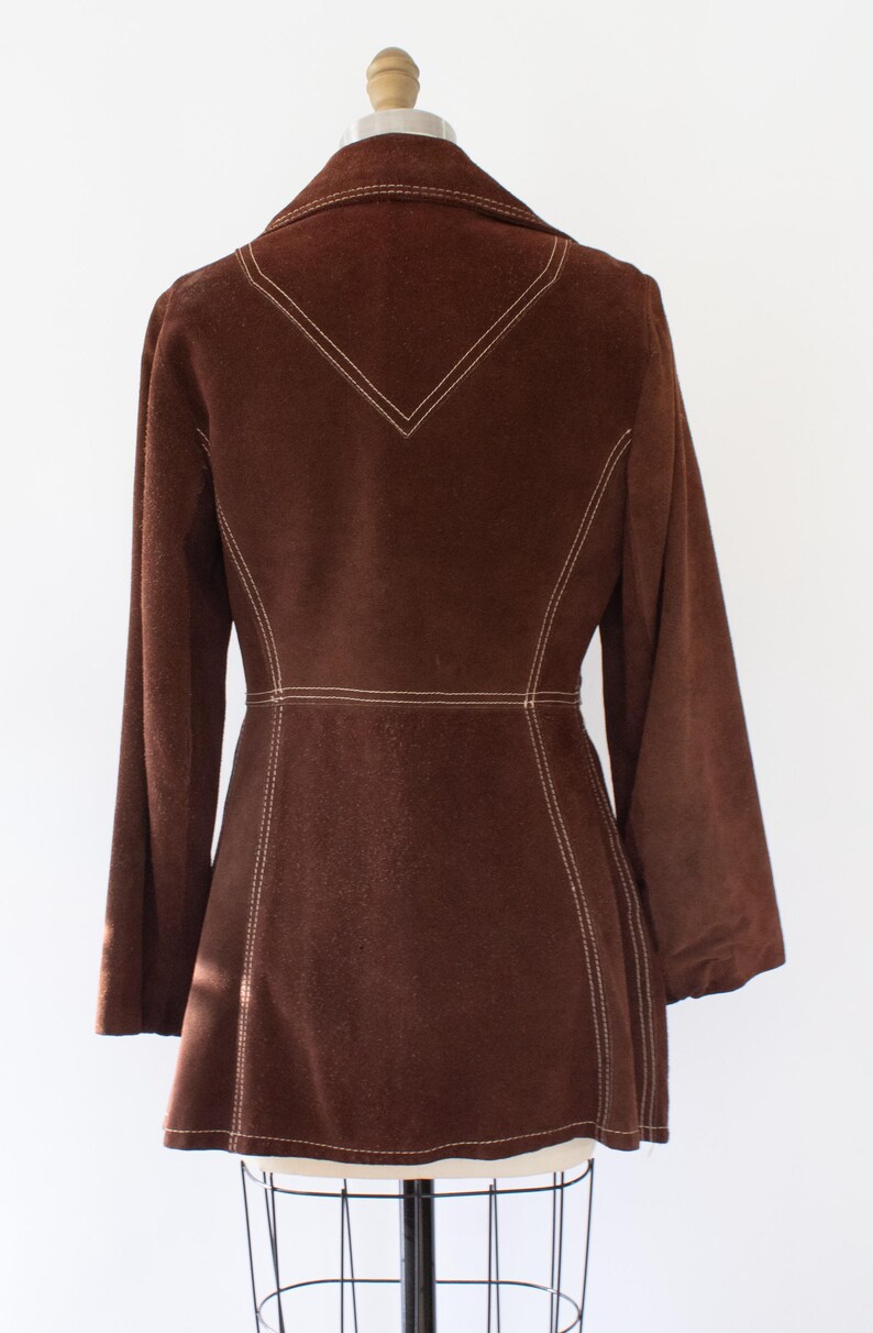 May include: A brown suede jacket with long sleeves and a tailored fit. The jacket features white stitching along the seams, collar, and a decorative V-shape on the back. The jacket is displayed on a mannequin.