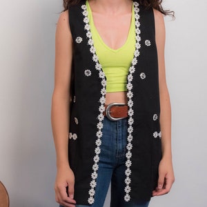 May include: A black sleeveless vest with white daisy-shaped trim and silver buttons. The vest has a long, flowing silhouette and is worn over a green tank top and blue jeans.