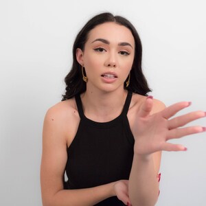 May include: A woman with long dark hair wearing a black tank top and gold earrings is talking and gesturing with her right hand.