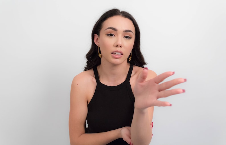 May include: A woman with long dark hair wearing a black tank top and gold earrings is talking and gesturing with her right hand.