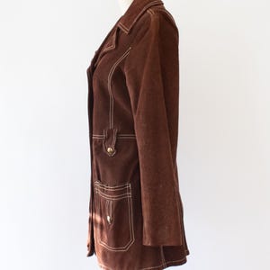 May include: A brown suede jacket with white stitching details. The jacket features a notched collar, two front pockets, and gold-tone buttons. The jacket is displayed on a mannequin.