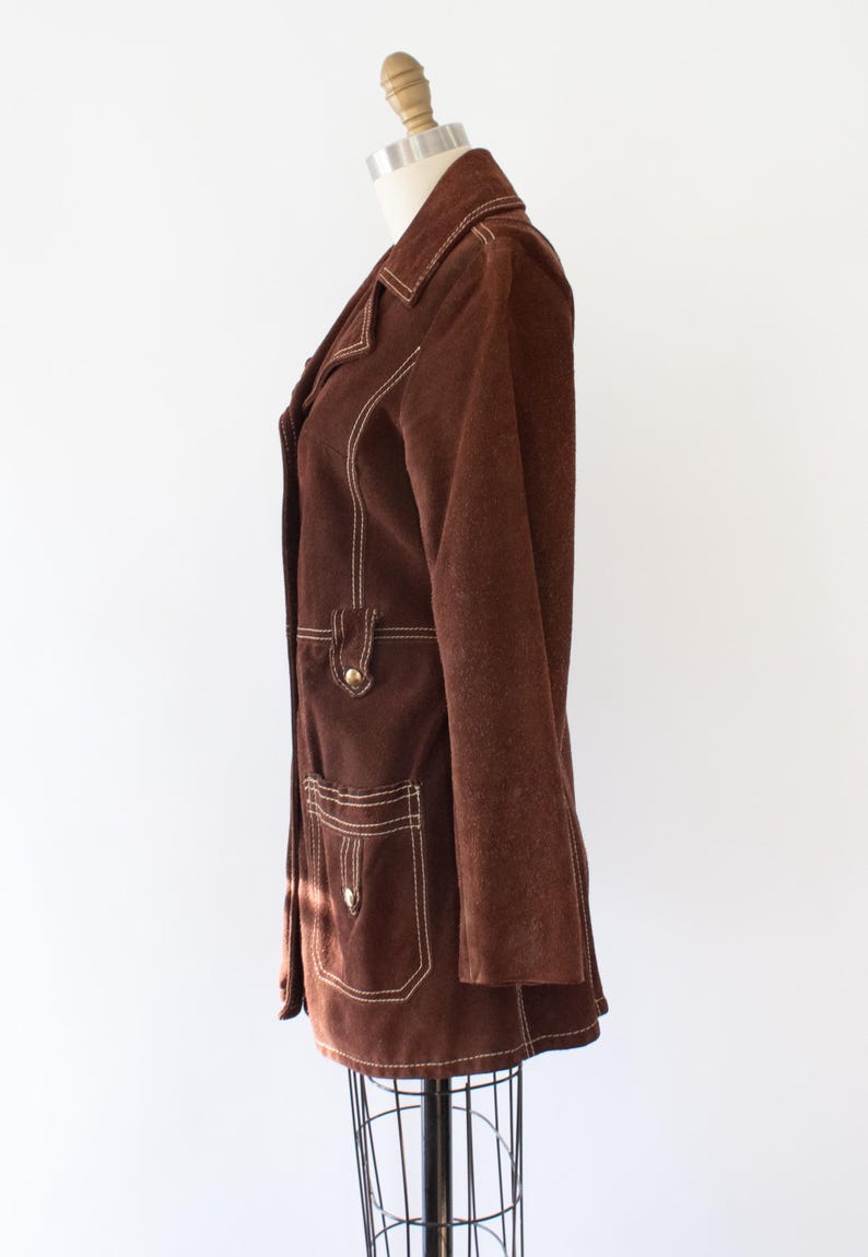 May include: A brown suede jacket with white stitching details. The jacket features a notched collar, two front pockets, and gold-tone buttons. The jacket is displayed on a mannequin.