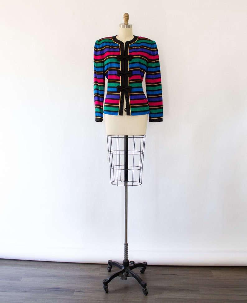 May include: A striped blazer with a black background and colorful stripes in pink, blue, green, and yellow. The blazer has black bows on the front.