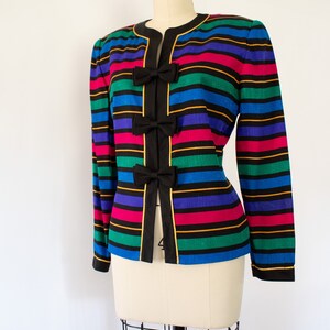 May include: A striped blazer with a black background and colorful stripes of blue, green, pink, and purple. The blazer has three black bows with gold trim on the front.