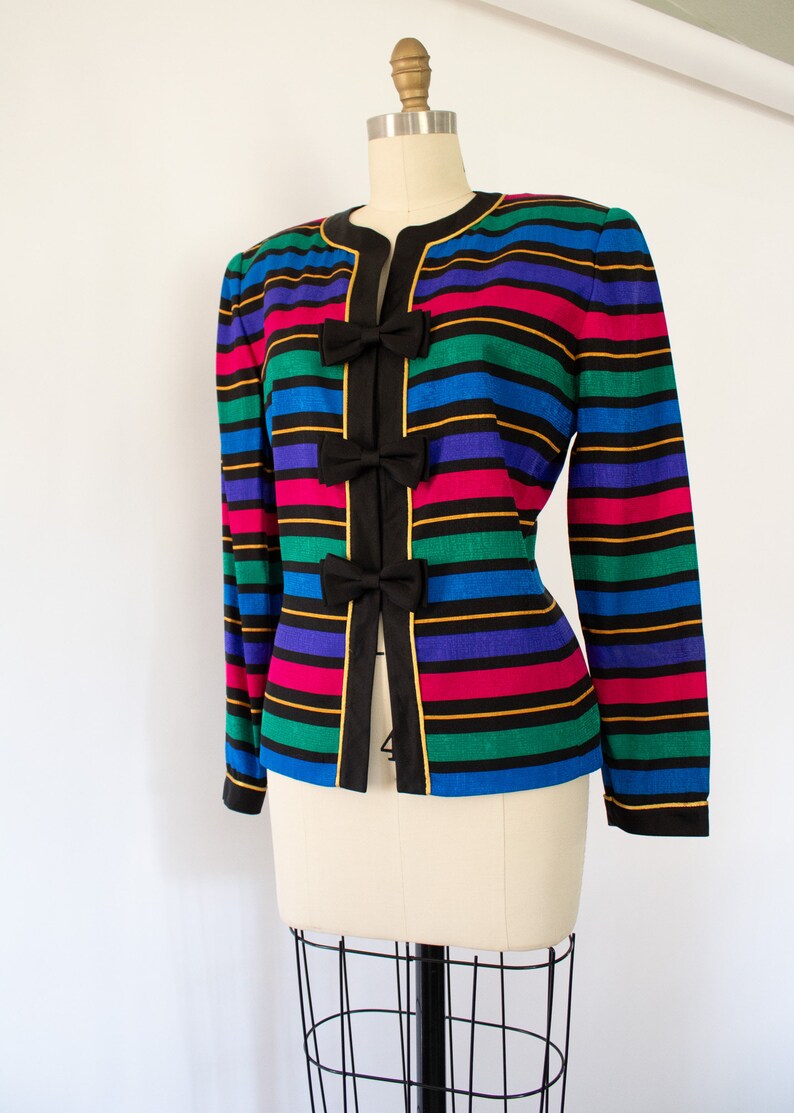 May include: A striped blazer with a black background and colorful stripes of blue, green, pink, and purple. The blazer has three black bows with gold trim on the front.