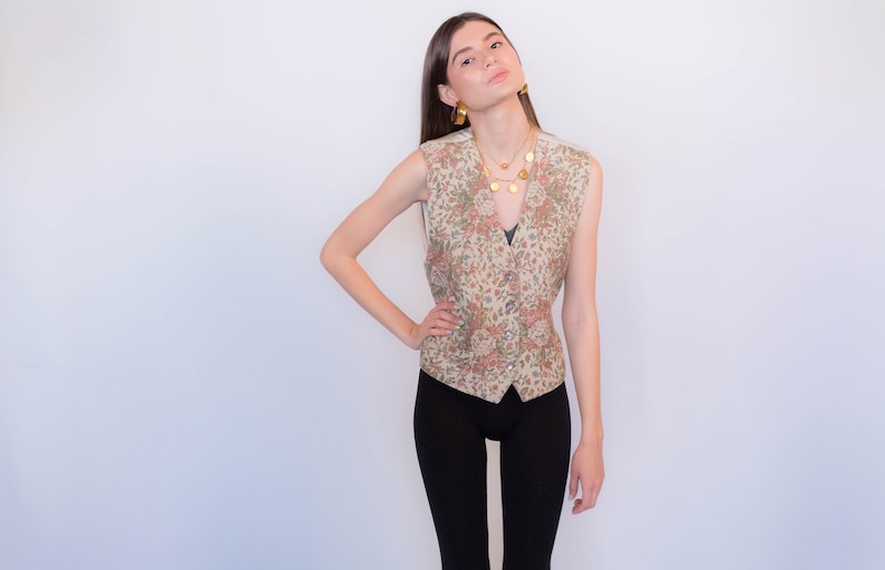 May include: A sleeveless floral patterned vest with a button closure. The vest is beige with pink and green floral accents. The model is wearing black pants and gold jewelry.