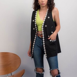 May include: A black sleeveless vest with white floral trim and large buttons. The vest is worn over a lime green tank top and blue jeans with distressed knees.