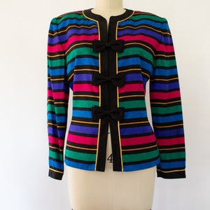 May include: A colorful striped blazer with black bows down the front. The blazer has a black collar and gold trim. The stripes are in shades of blue, green, pink, and yellow.