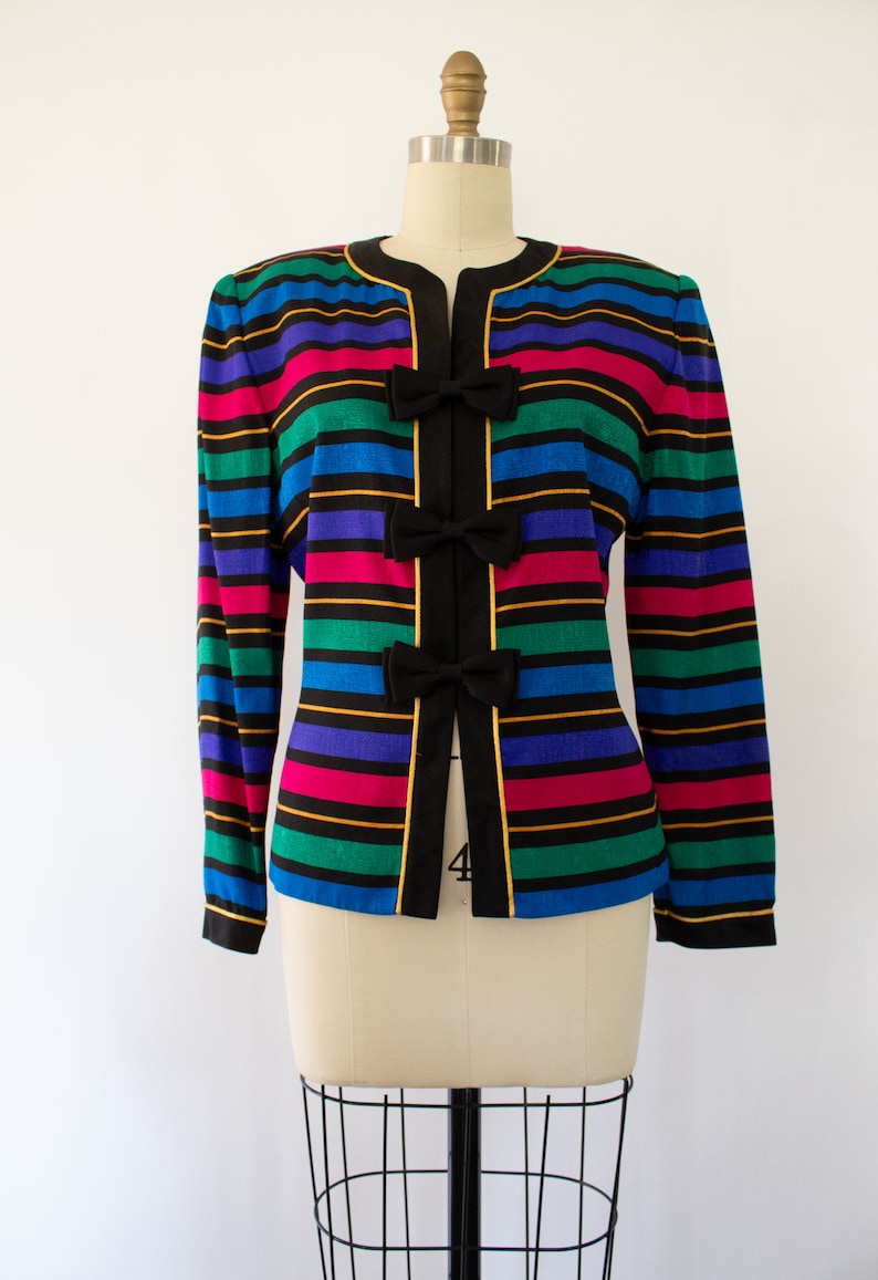 May include: A colorful striped blazer with black bows down the front. The blazer has a black collar and gold trim. The stripes are in shades of blue, green, pink, and yellow.