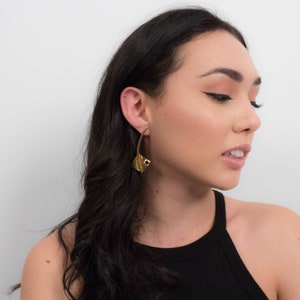 May include: A woman with long black hair wearing a black tank top and gold earrings. The earrings are large and have a geometric design.