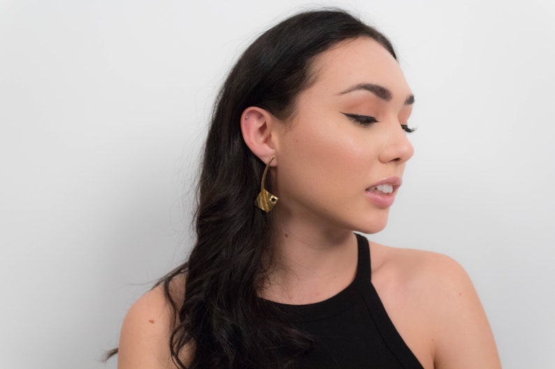 May include: A woman with long black hair wearing a black tank top and gold earrings. The earrings are large and have a geometric design.