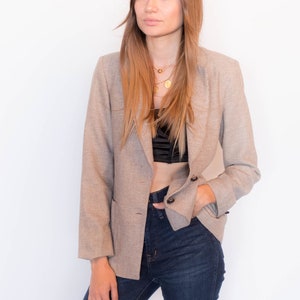 Puede incluir: Una mujer con un blazer beige sobre un top negro corto y unos vaqueros azules oscuros. Lleva un collar de oro con un colgante.