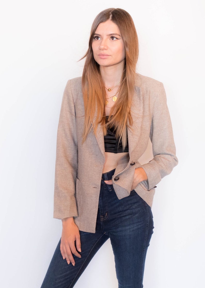 Puede incluir: Una mujer con un blazer beige sobre un top negro corto y unos vaqueros azules oscuros. Lleva un collar de oro con un colgante.