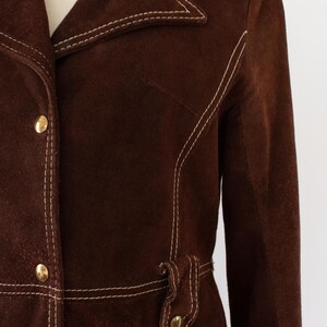 May include: A brown suede jacket with white stitching and gold buttons. The jacket features a notched collar, a belt detail at the waist, and a tailored fit. This vintage-inspired jacket is a stylish outerwear option.