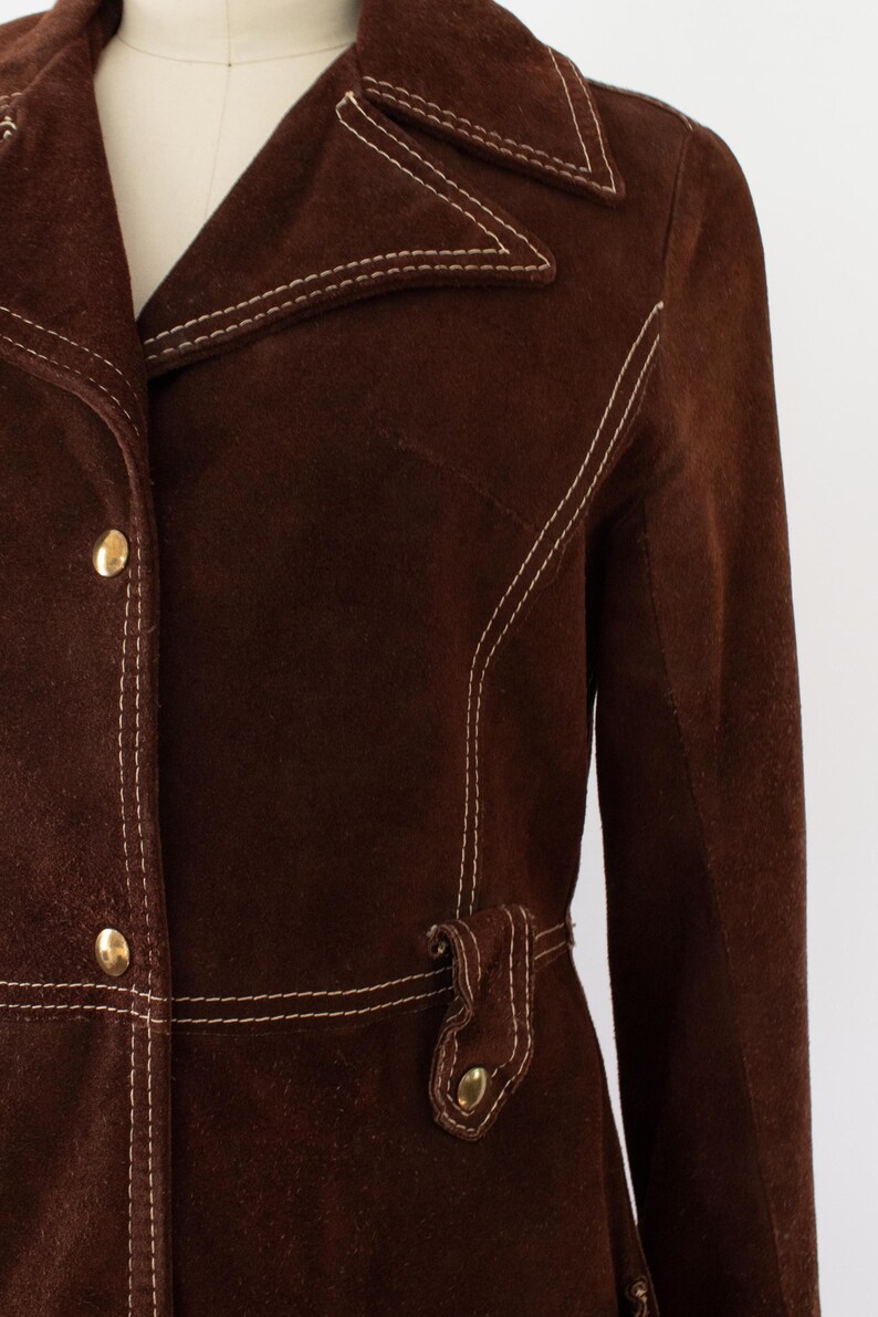 May include: A brown suede jacket with white stitching and gold buttons. The jacket features a notched collar, a belt detail at the waist, and a tailored fit. This vintage-inspired jacket is a stylish outerwear option.