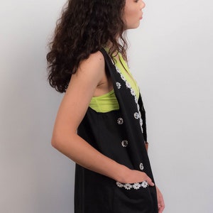 May include: A black sleeveless vest with white daisy-shaped embellishments. The vest has a V-neck and pockets with white daisy trim. The model is wearing a green tank top underneath.
