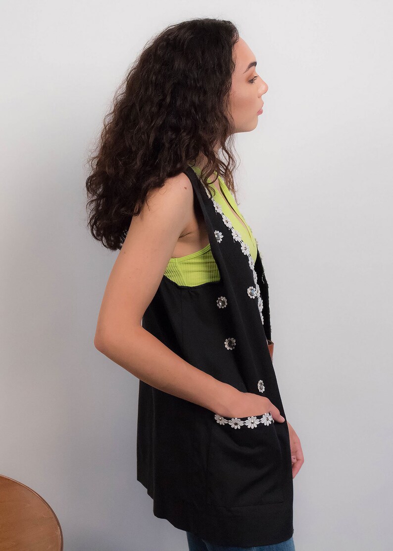 May include: A black sleeveless vest with white daisy-shaped embellishments. The vest has a V-neck and pockets with white daisy trim. The model is wearing a green tank top underneath.