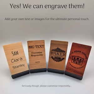 May include: Four wooden engraved phone stands with black bases. The stands feature custom text and logos, including "SAM Clara Stanley", "BIG TEXT Customize Responsibly", "BAKER HARDWOODS", and "THE HOME OFFICE co-working community". The image includes the text "Yes! We can engrave them!"