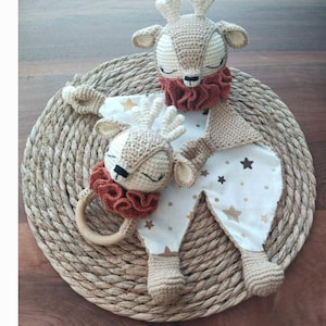 May include: A handmade crochet deer baby toy and rattle set. The deer features a beige body, cream antlers, and a rust-colored ruffle. The rattle has a wooden ring. The set is on a white and gold star patterned fabric.