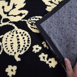 Könnte beinhalten: Schwarz-cremefarbener Teppich mit floralem Muster. Der Teppich hat ein sich wiederholendes Muster aus stilisierten Blumen und Blättern. Die Rückseite des Teppichs ist grau mit einer rutschfesten Textur. Der Teppich ist rechteckig.