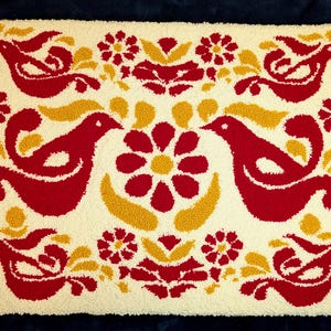 Könnte beinhalten: Cremefarbener Teppich mit einem sich wiederholenden Muster aus roten Vögeln, Blumen und gelben Akzenten. Das Design ist symmetrisch, mit sich zugewandten Vögeln. Der Teppich hat ein strukturiertes, getuftetes Aussehen, vor dunklem blauem Hintergrund.