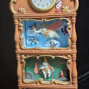 Puede incluir: Un reloj decorativo con el tema de Dumbo. El reloj tiene un marco de madera con detalles dorados y una esfera redonda. El reloj también presenta a Dumbo y otros personajes de la película en los diferentes niveles.