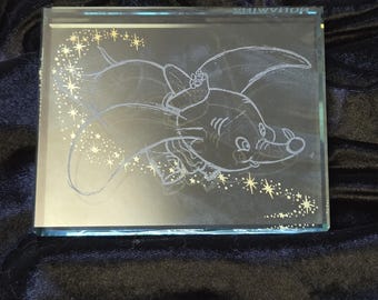 May include: A rectangular glass block featuring an etched image of Dumbo the elephant, surrounded by stars. The artwork is in a light color, set against a dark background. The name "MOUHAMIDA" is etched on the top edge.