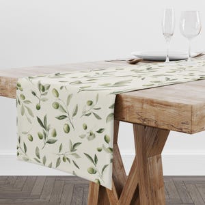 May include: A cream-colored table runner featuring a repeating watercolor pattern of green olive branches with leaves and olives. The runner is draped over a wooden table, with two clear glass wine glasses and a white plate visible nearby.