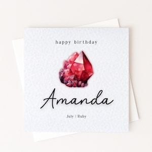 May include: A square greeting card with a textured white surface featuring a watercolour illustration of a red ruby crystal. The text reads happy birthday, the name Amanda in black script, and the words July | Ruby at the bottom of the card.
