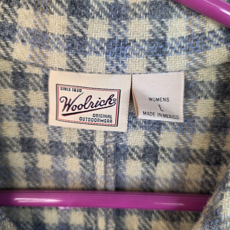 May include: A blue and cream plaid wool-blend shirt featuring a white interior tag with the text Woolrich Since 1830 Original Outdoorwear and a second tag indicating size L and Made in Mexico, hanging on a bright purple plastic clothes hanger.