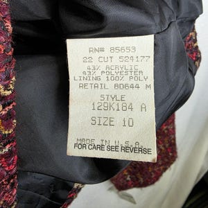 May include: Close-up of a burgundy tweed jacket lining with a tag. The tag reads "RN# 85653, SIZE 10, MADE IN U.S.A." The jacket's fabric is a mix of red, gold, and black threads.