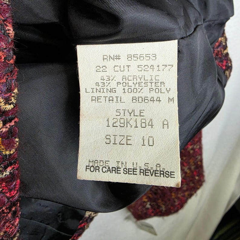 May include: Close-up of a burgundy tweed jacket lining with a tag. The tag reads "RN# 85653, SIZE 10, MADE IN U.S.A." The jacket's fabric is a mix of red, gold, and black threads.