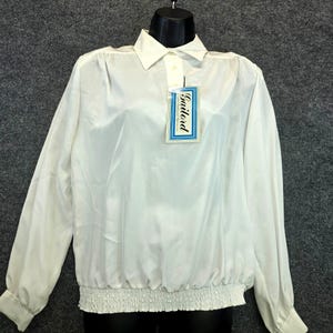 May include: A white long-sleeve blouse with a collared neckline and a gathered waist. The blouse has a button closure at the collar and cuffs. A tag with the name "Gailord" is attached. The fabric appears to be a lightweight, possibly silky material.
