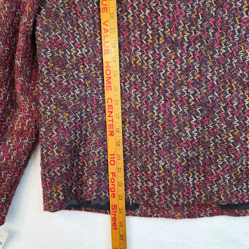 May include: A close-up of a burgundy tweed jacket with a textured, woven pattern. The jacket features a mix of colors including red, yellow, and purple. The jacket has a black lining and a measurement tool is placed next to the jacket.