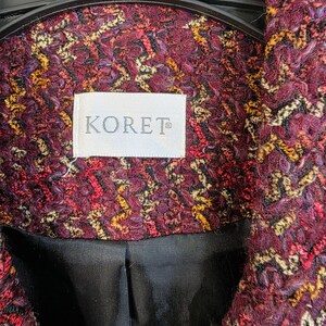 May include: Close-up of a burgundy jacket with a textured, woven pattern in shades of red, yellow, and black. The jacket features a label that reads "KORET" in white lettering. The lining is black.