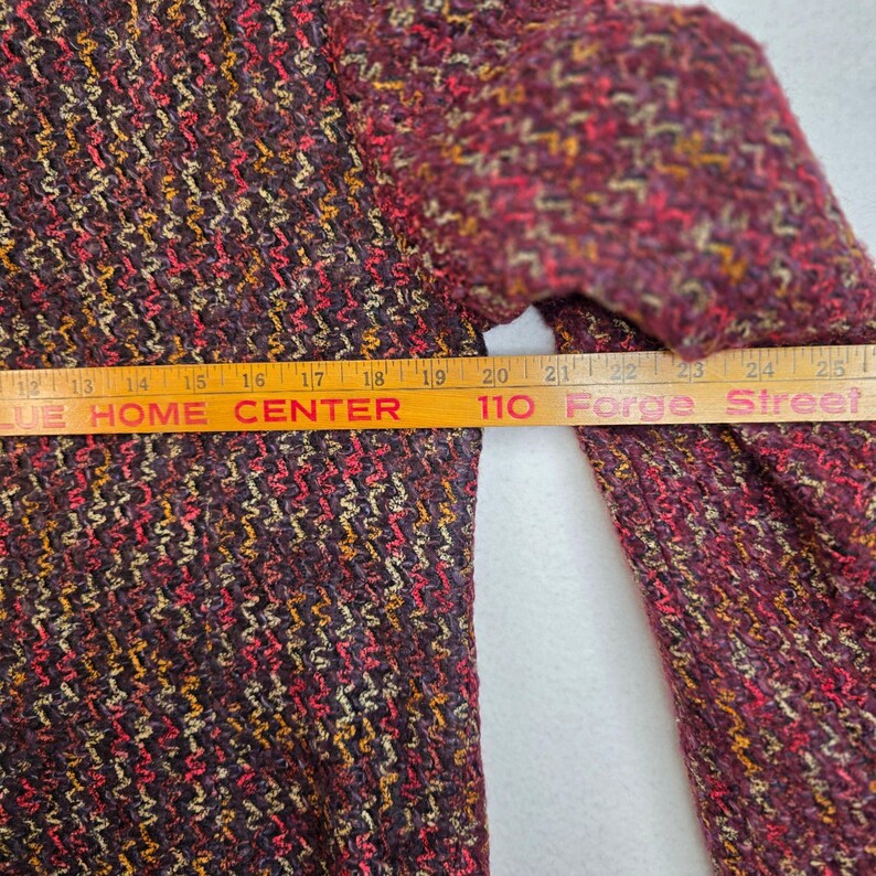 May include: Close-up of a textured, multi-colored jacket in shades of burgundy, red, and gold. The jacket's fabric has a woven appearance. A measuring tape is placed across the jacket, with the text "BLUE HOME CENTER" and "110 Forge Street" visible.