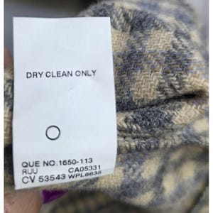 May include: A white fabric care label attached to a textured beige and gray plaid garment. The label features the text DRY CLEAN ONLY, a circle symbol, and the codes QUE NO.1650-113, RUU, CA05331, CV 53543, and WPL6635 printed in black ink.