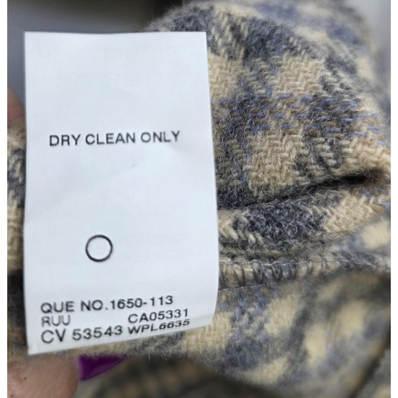 May include: A white fabric care label attached to a textured beige and gray plaid garment. The label features the text DRY CLEAN ONLY, a circle symbol, and the codes QUE NO.1650-113, RUU, CA05331, CV 53543, and WPL6635 printed in black ink.