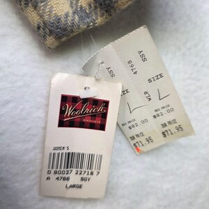 May include: A beige and grey plaid fabric corner is attached to two white paper tags. One tag features the Woolrich logo with a red and black buffalo check background and the text 'LARGE'. The second tag displays size L and product codes.
