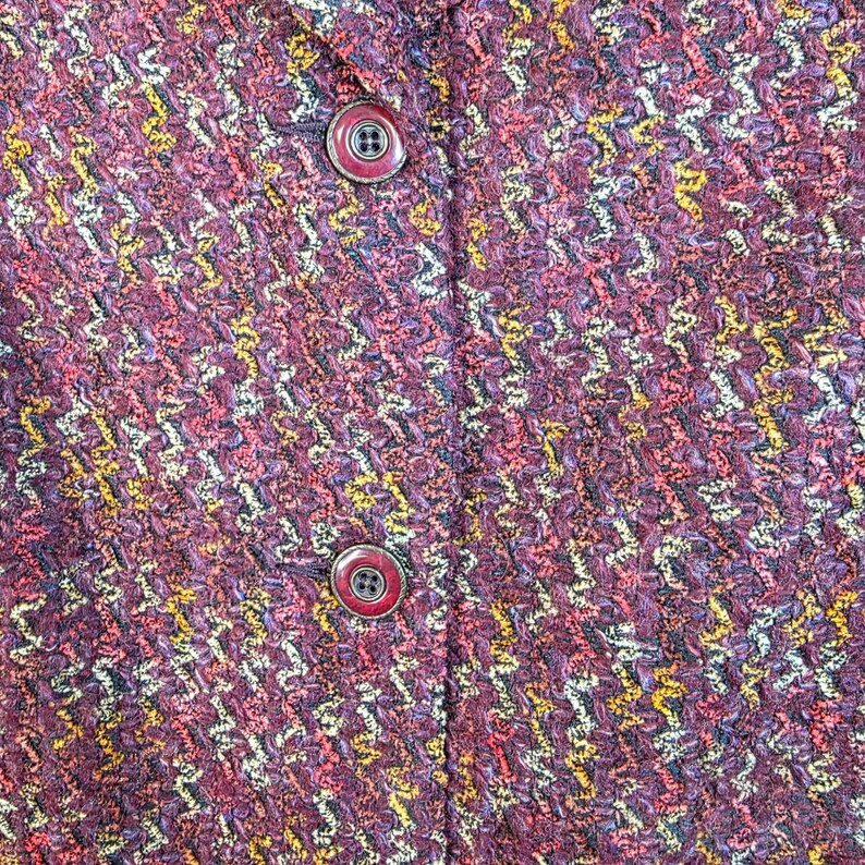 May include: Close-up of a textured, multi-colored fabric with a woven pattern. The fabric is primarily burgundy with accents of yellow, pink, and white. Two round buttons with red rims are visible.