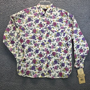 May include: A button-down shirt with a white background and a floral pattern in shades of purple, pink, and yellow. The long-sleeved shirt has a collar and a pocket on the left chest. A tag is attached to the shirt.